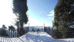 Un terrazzo innevato con due sedie e un tavolo, circondato da ringhiere metalliche e alberi, con vista su un corpo d'acqua.