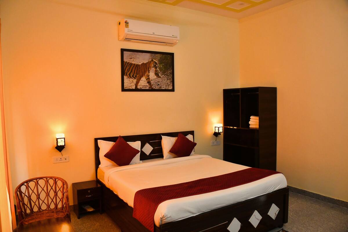 A hotel room with a bed, a nightstand, a chair, a wall-mounted air conditioner, a wall-mounted light, and a framed picture of a zebra.
