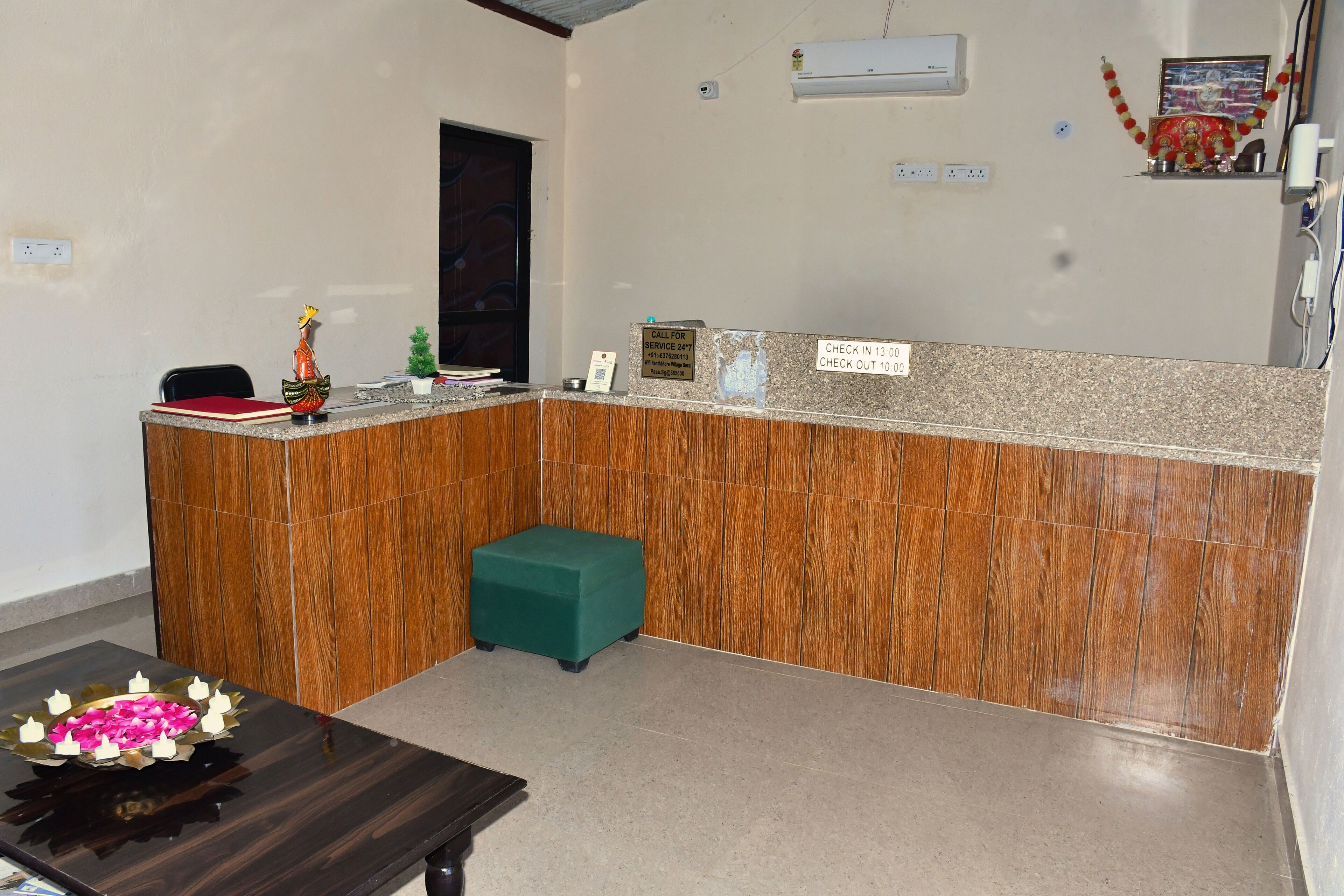 A reception area with a wooden counter, a green stool, and a small statue. There are signs indicating check-in and check-out times, and an air conditioning unit on the wall.