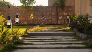 A stone pathway leading to a building, flanked by greenery and lanterns. - Ranthambore Village Serai (Sawai Madhopur)