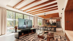 A modern living room with a large TV, wooden ceiling beams, a dining area, and a kitchen in the background. - The Urban Retreat Villa Tiga - 4BR Villa, Seminyak with Butler, cook and gym (Kabupaten Badung)