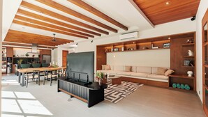 A modern living room with a wooden ceiling, a large TV, a sofa, and a dining area with a kitchen in the background.