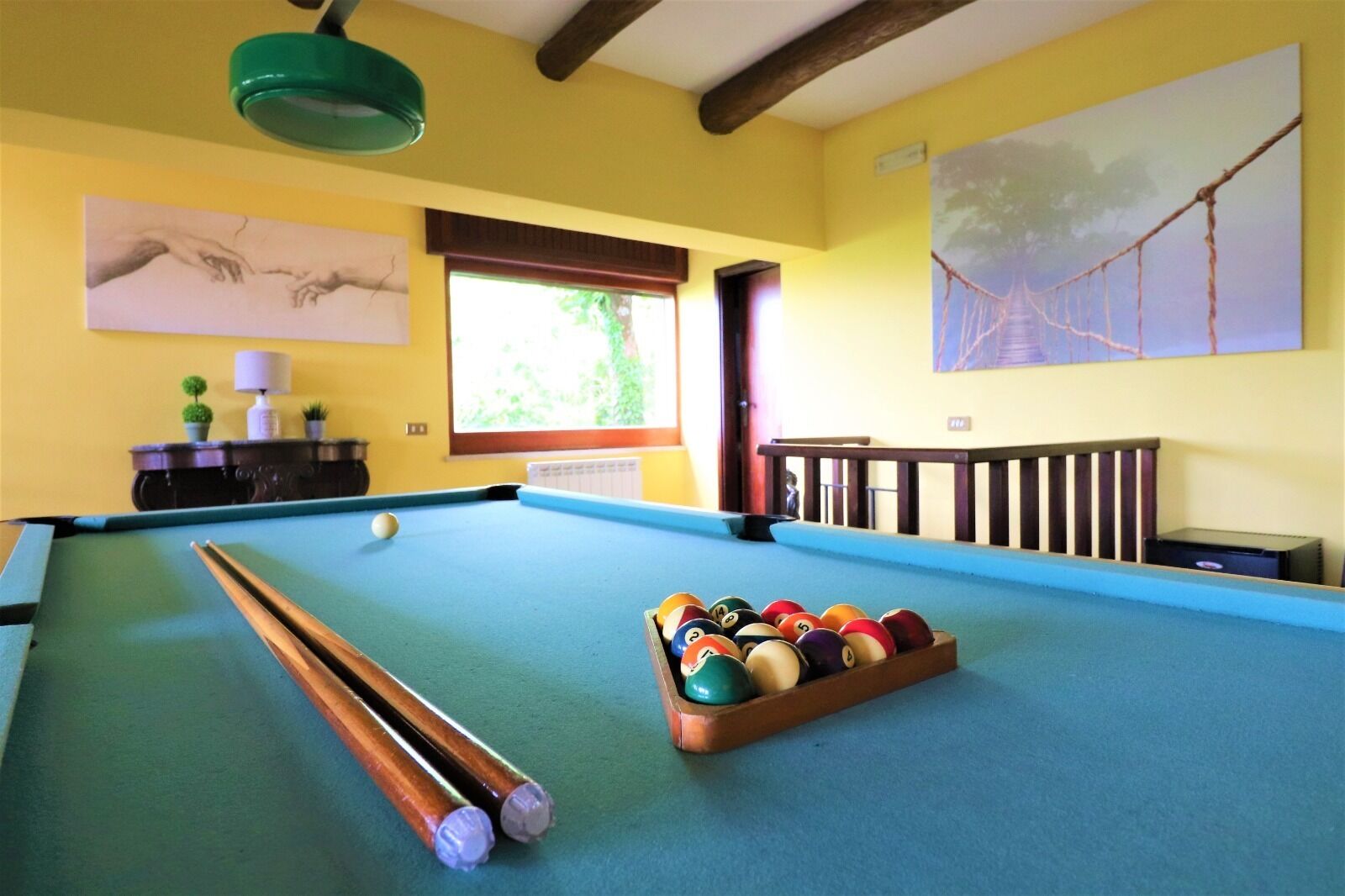 A pool table with balls and cues, a window with a view of greenery, and a painting on the wall.