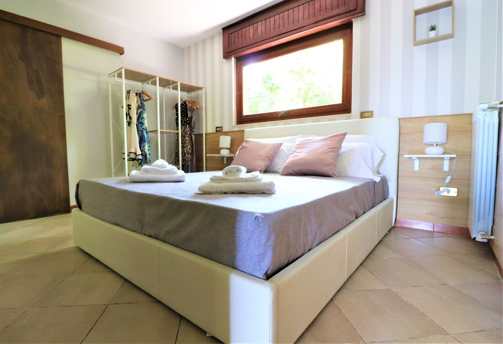 Un dormitorio con una cama grande, un armario, una ventana y una mesita de noche con lámpara.