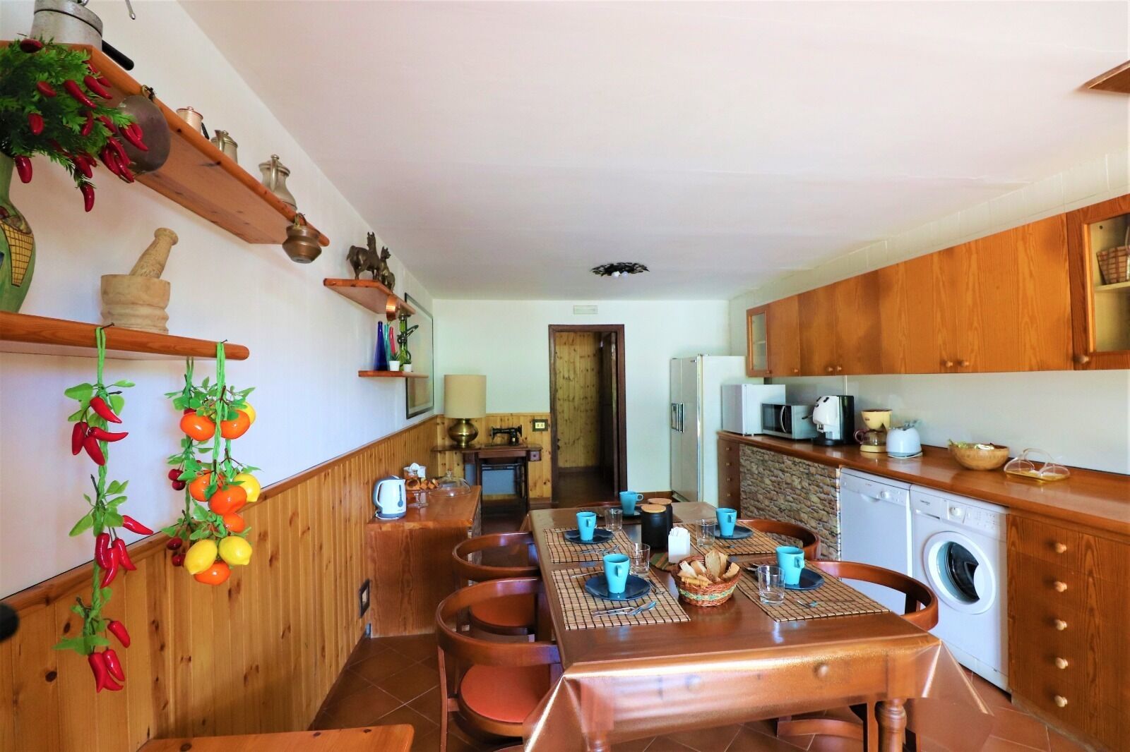 A cozy kitchen with wooden cabinets, a dining table set with blue cups, and a hanging plant with red peppers.