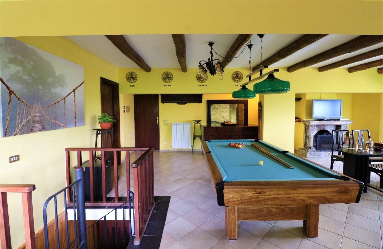 A pool table with a billiard cue, a television, and a fireplace in a room with yellow walls and wooden beams.