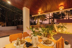 An outdoor dining area with wooden tables and chairs, a vase with flowers, and a view of palm trees and a sunset. - ELIVAAS House of Neptune 1 BHK Boutique (Morjim)