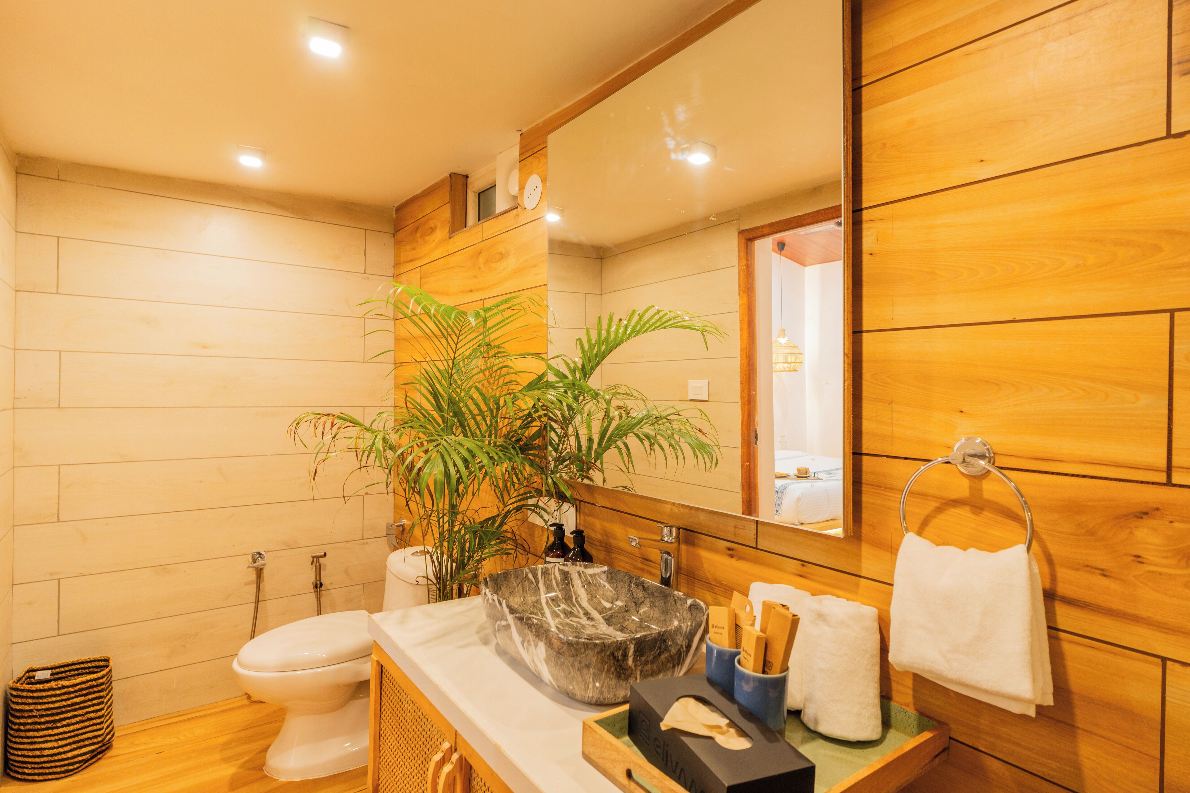 A modern bathroom with a wooden vanity, a large mirror, a sink with a marble basin, a toilet, and a potted plant.