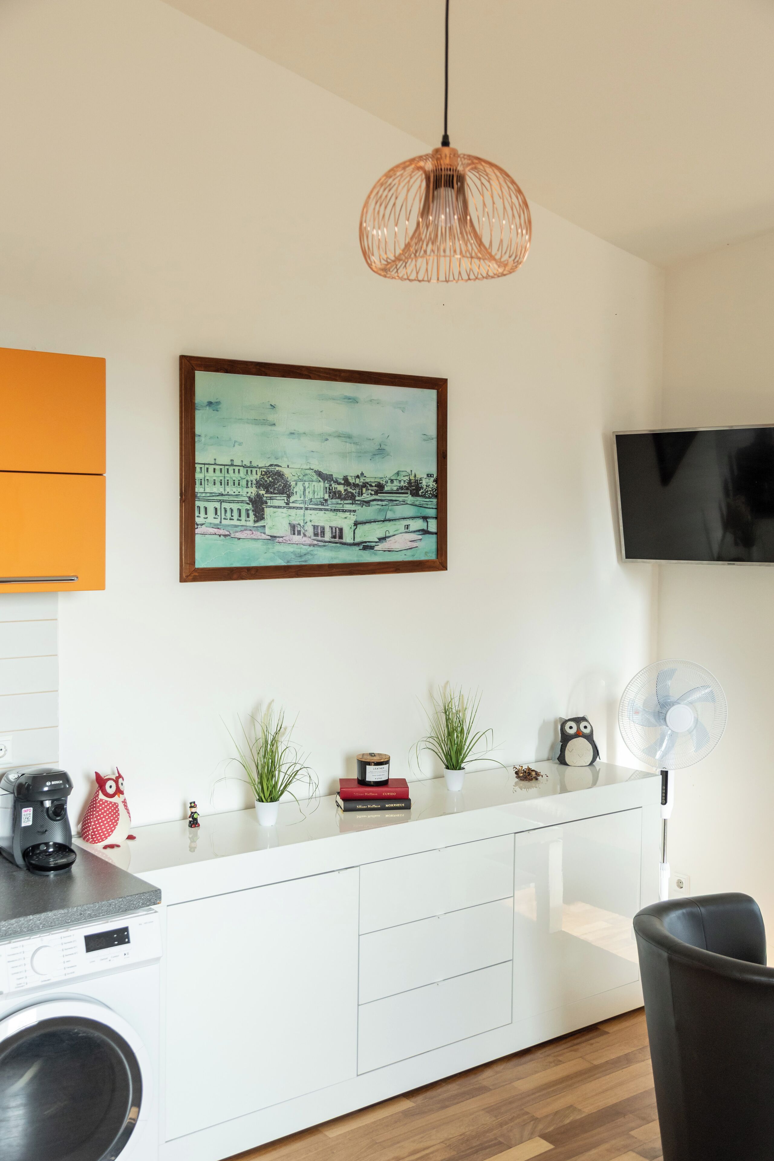 A modern kitchen with a white countertop, a hanging pendant light, a framed cityscape painting, a television mounted on the wall, and a small fan on the counter.