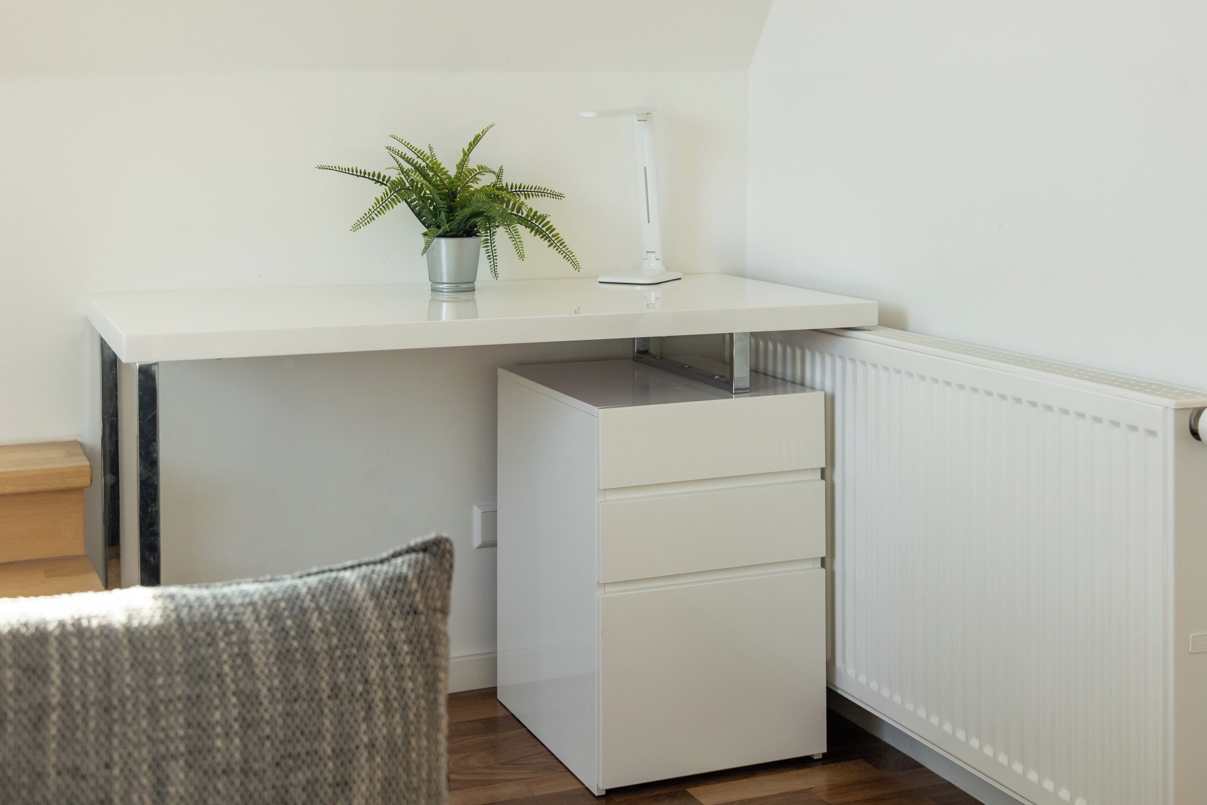 Un bureau compact et blanc, agrémenté d’une plante et d’une lampe de bureau, une chaise et un radiateur, dans une petite pièce.