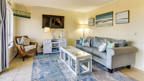 Una sala de estar con un sofá gris, una mesa de centro blanca, una silla de mimbre, una mesita auxiliar blanca y un tapete azul con estampado.