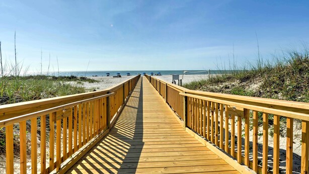 Un paseo marítimo de madera que conduce a una playa con dunas de arena y un cielo despejado.