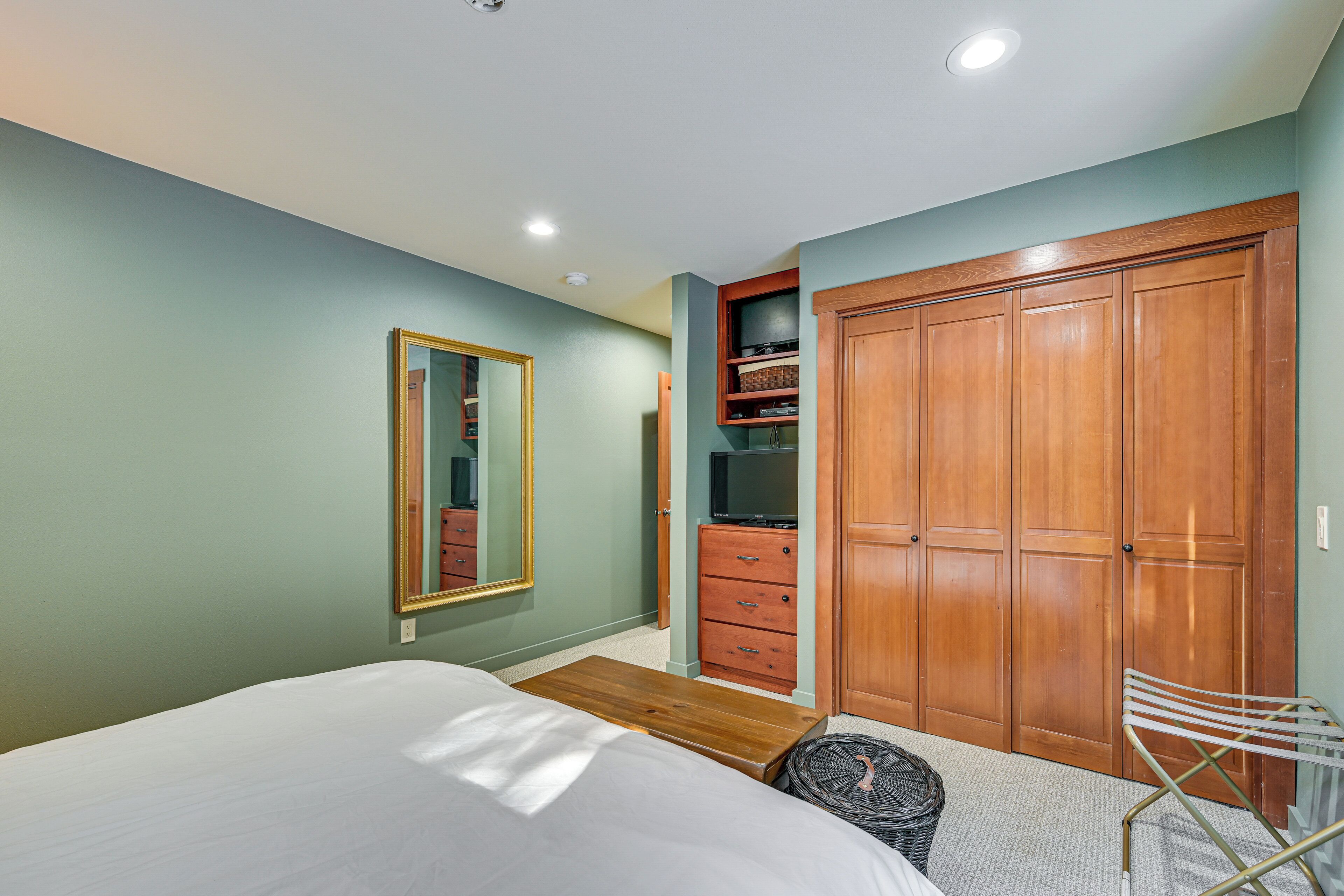 A bedroom with a bed, a wooden bench, a mirror, a wardrobe, and a shelf with a television.