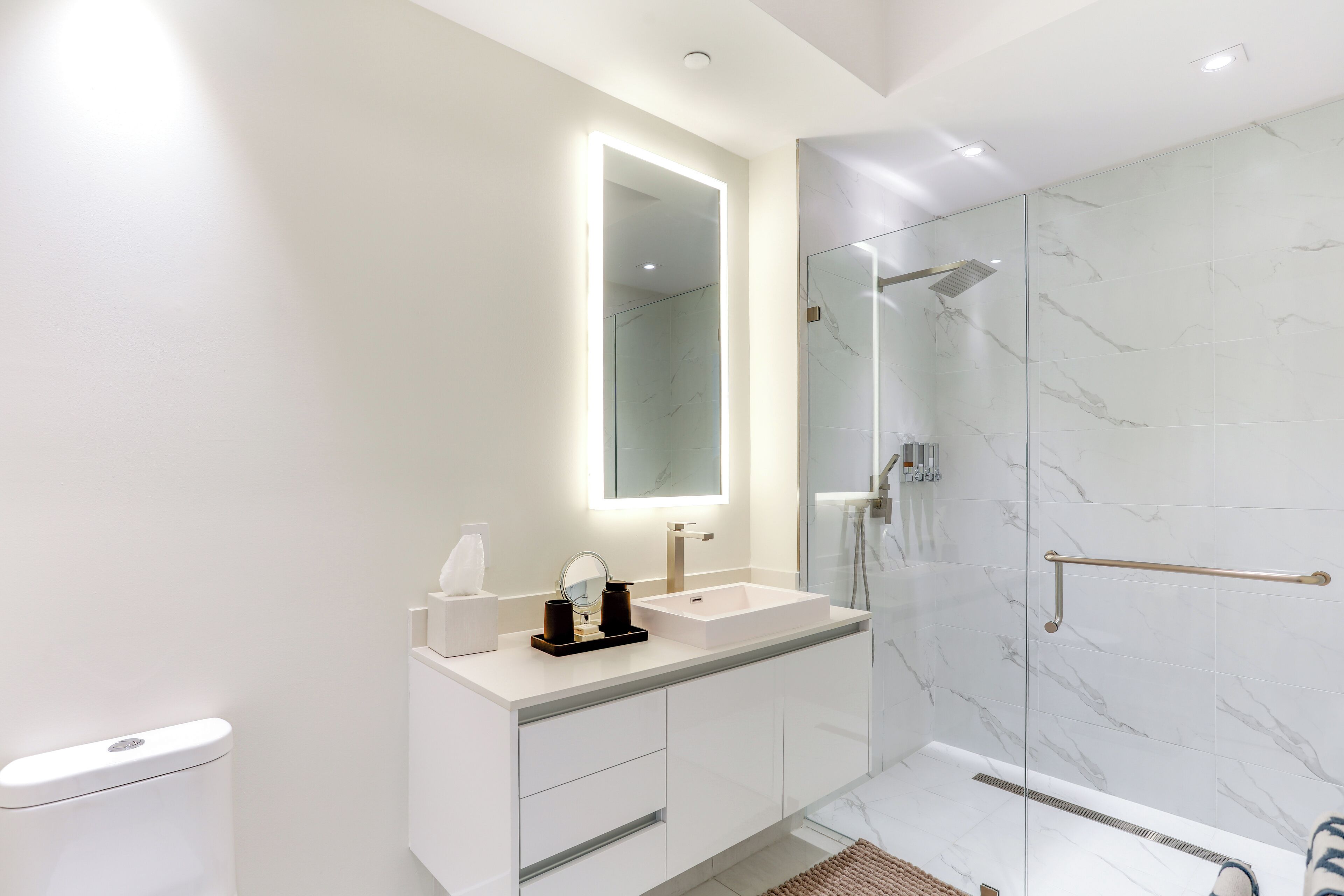 A modern bathroom with a white vanity, a large mirror, a glass shower enclosure, and marble walls.