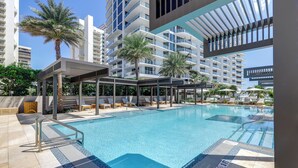 A swimming pool with lounge chairs and palm trees, surrounded by modern buildings. - Condo at Amrit Ocean Resort w/ Panoramic Views (Singer Island)