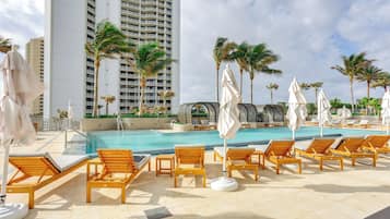A poolside area with lounge chairs and umbrellas, a large pool, and tall buildings in the background.