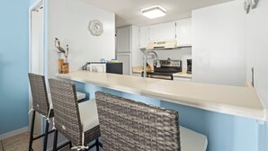 A compact kitchen with white cabinets, a white countertop, and a built-in oven. There are two wicker chairs and a small dining table.