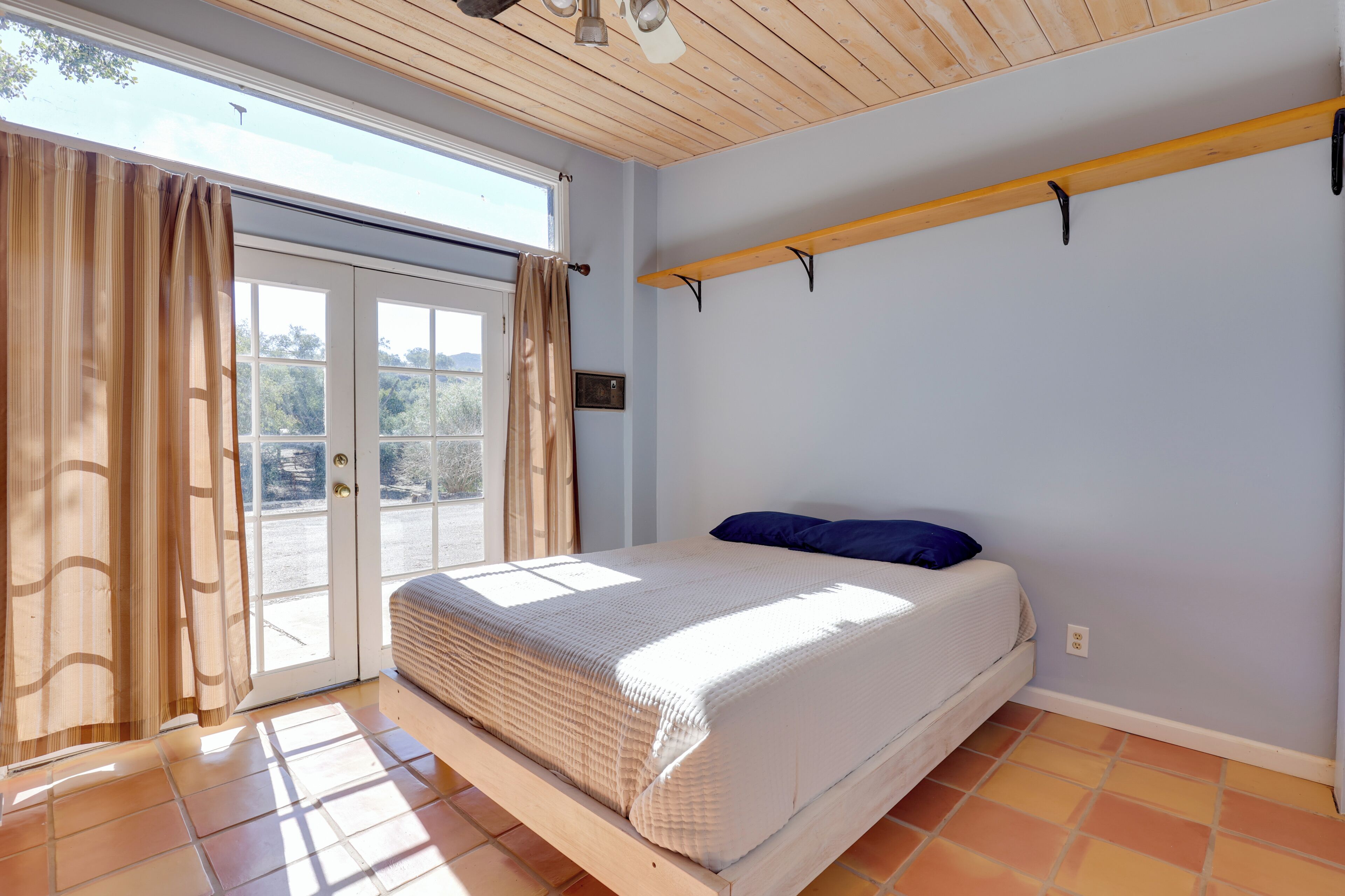 Ein Schlafzimmer mit einem Bett, einem Fenster mit Vorhängen, einer Holzbalkendecke und einem Fliesenboden.