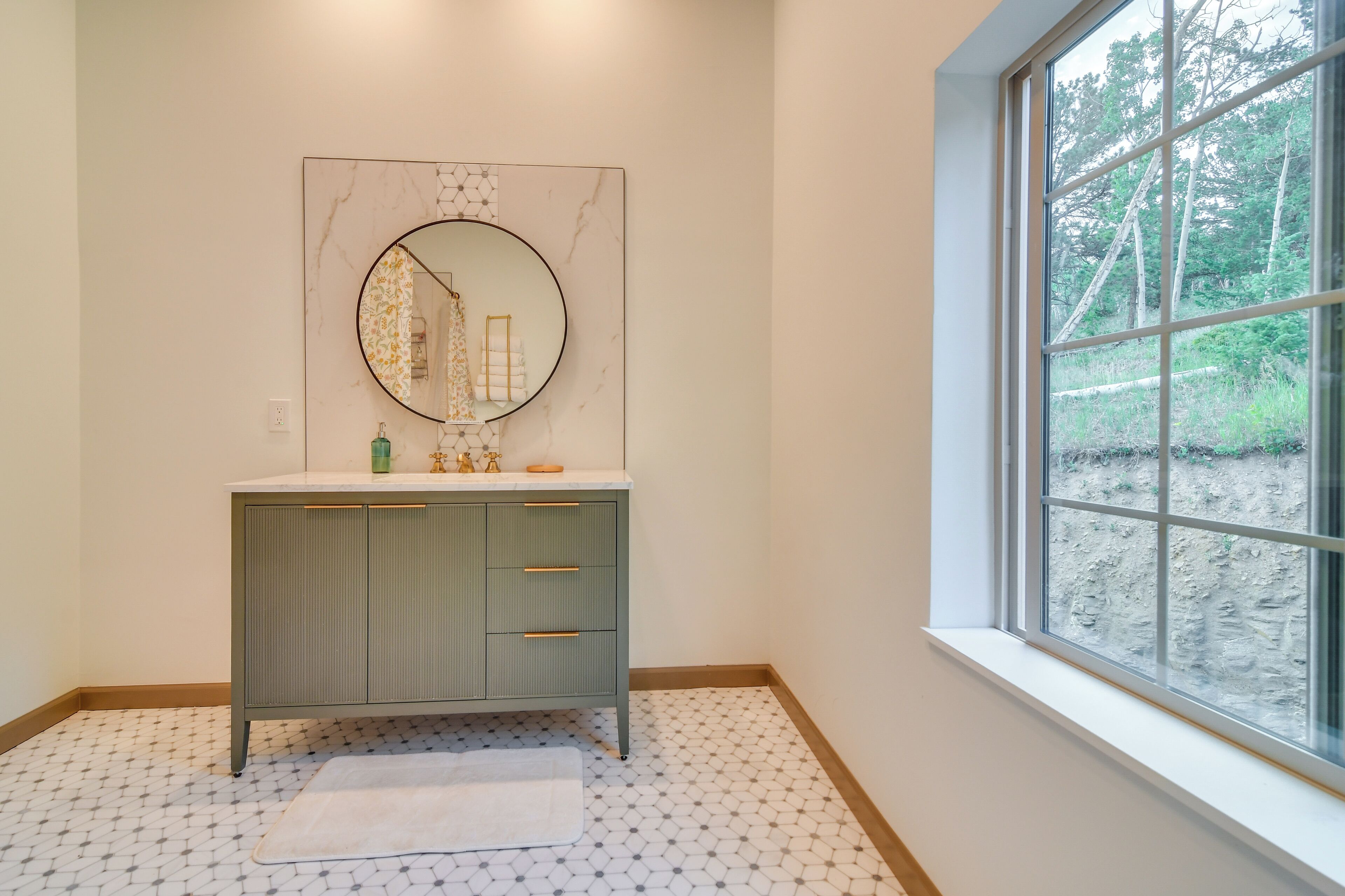 A bathroom with a marble vanity, a round mirror, a window with a view of trees, and a patterned floor.