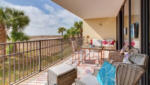 A balcony with wicker furniture, a table, and chairs, overlooking a beach with palm trees.