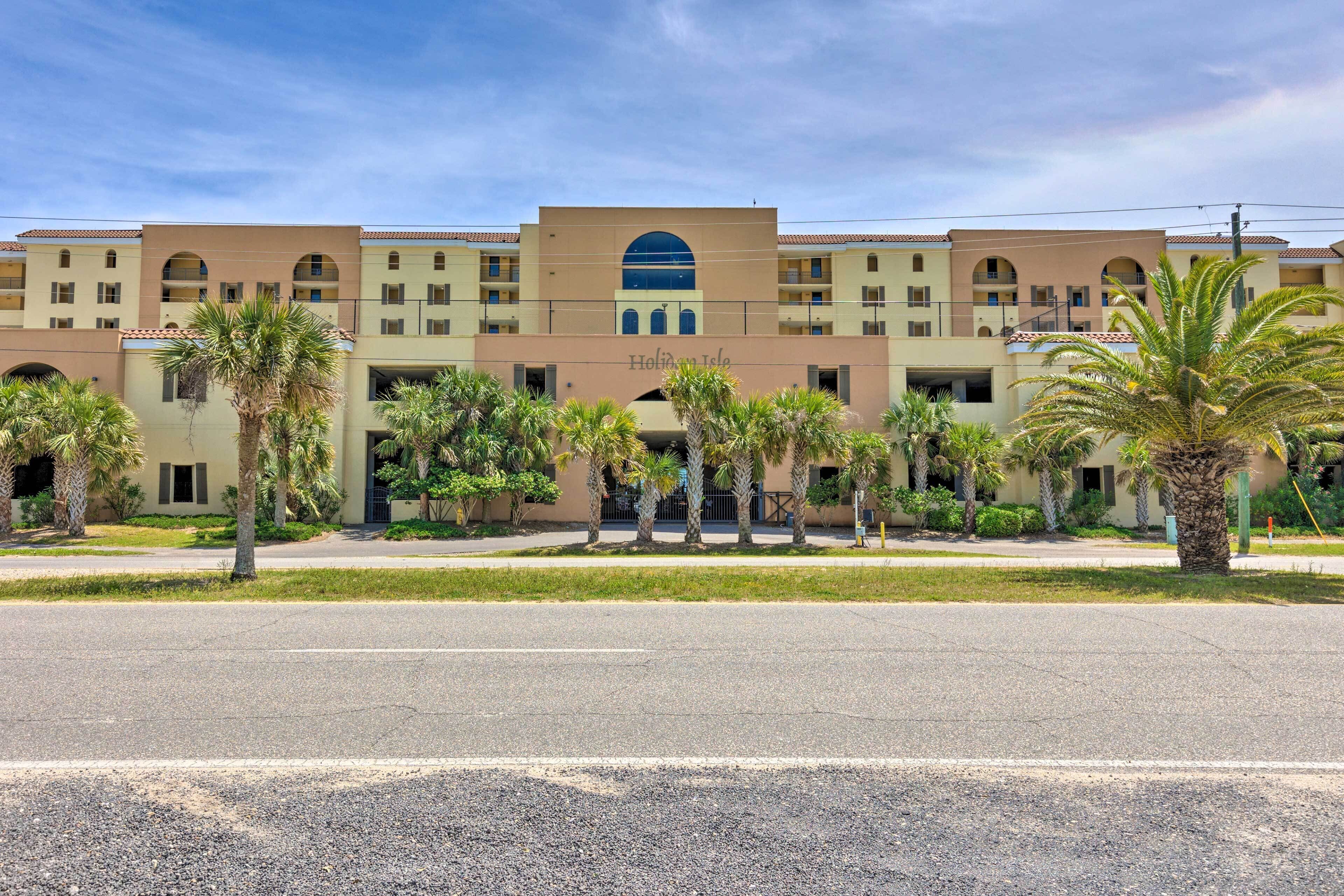 A hotel with a central arched entrance, multiple balconies, and palm trees in the foreground.