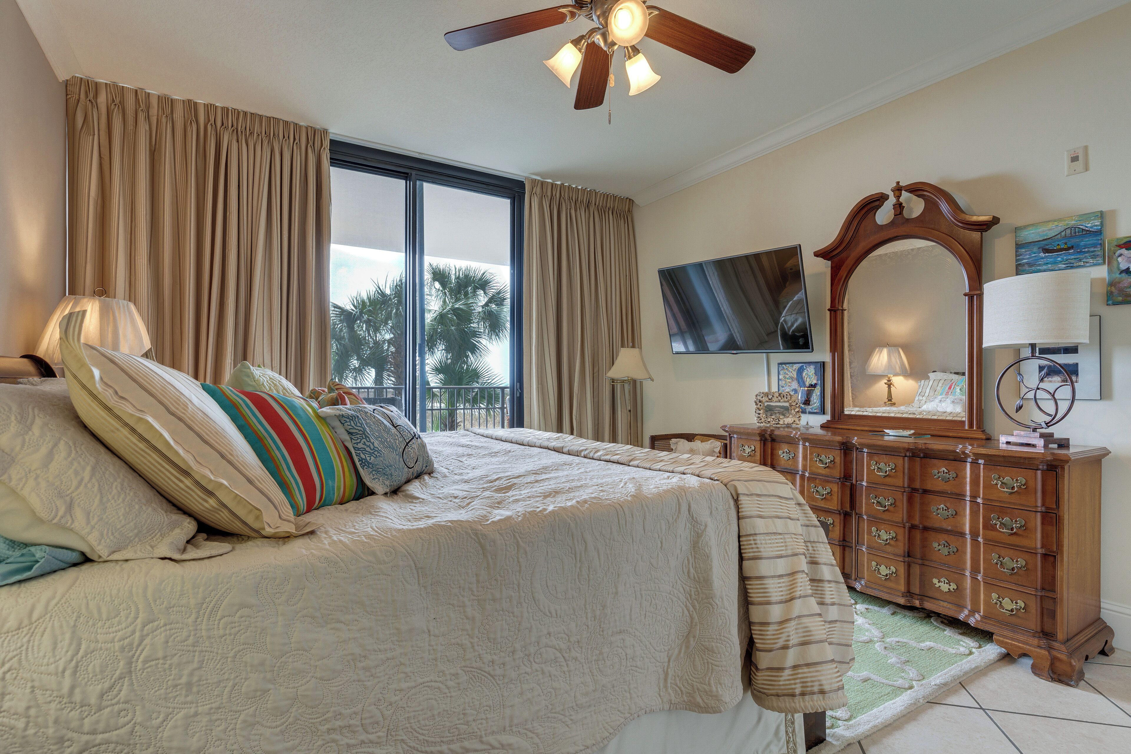 A bedroom with a large bed, a wooden dresser, a ceiling fan, a TV, and a window with curtains.