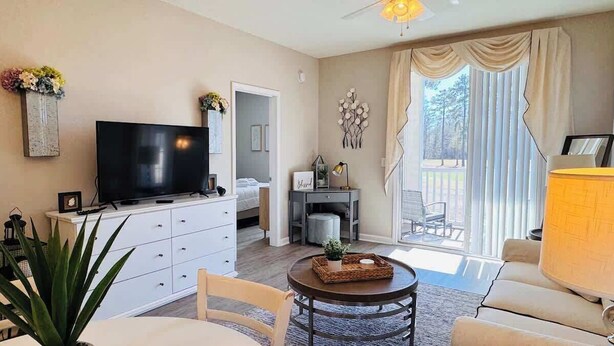 A living room with a TV, a dining table, and a sofa. There is a balcony with a view of trees. - River Oaks 2BR beautiful condo (Myrtle Beach)