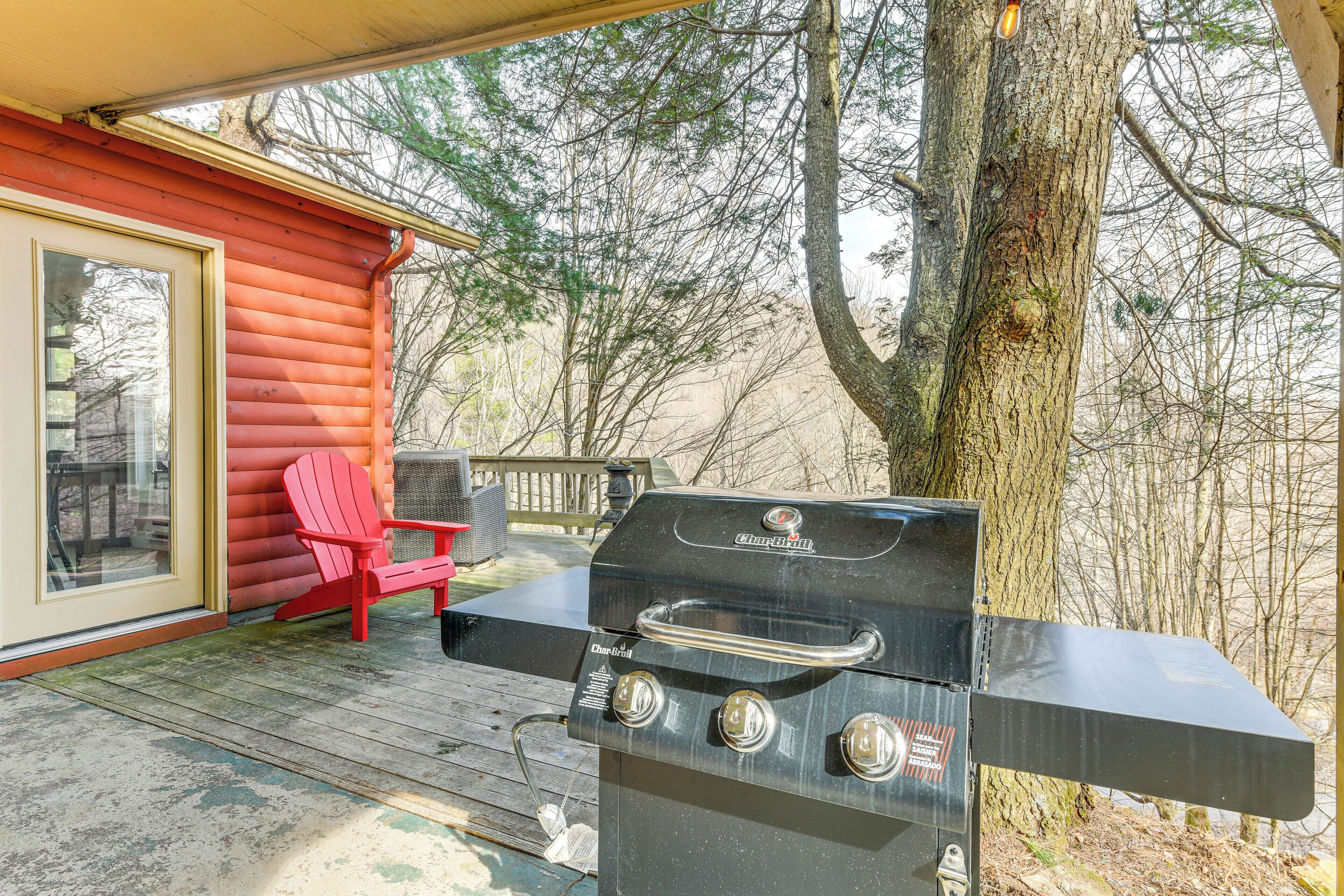 A barbecue grill on a wooden deck with a red chair and a glass door leading to a room.