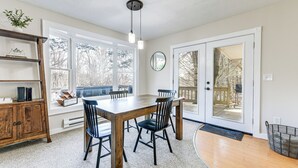 Un comedor con una mesa y sillas de madera, una estantería y un ventanal con vistas a árboles.