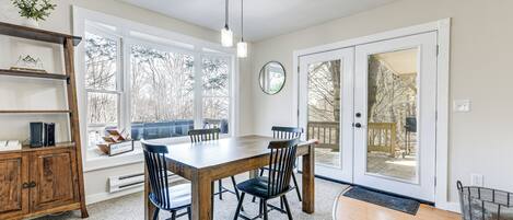 A dining area with a wooden table and chairs, a bookshelf, and a large window with a view of trees.