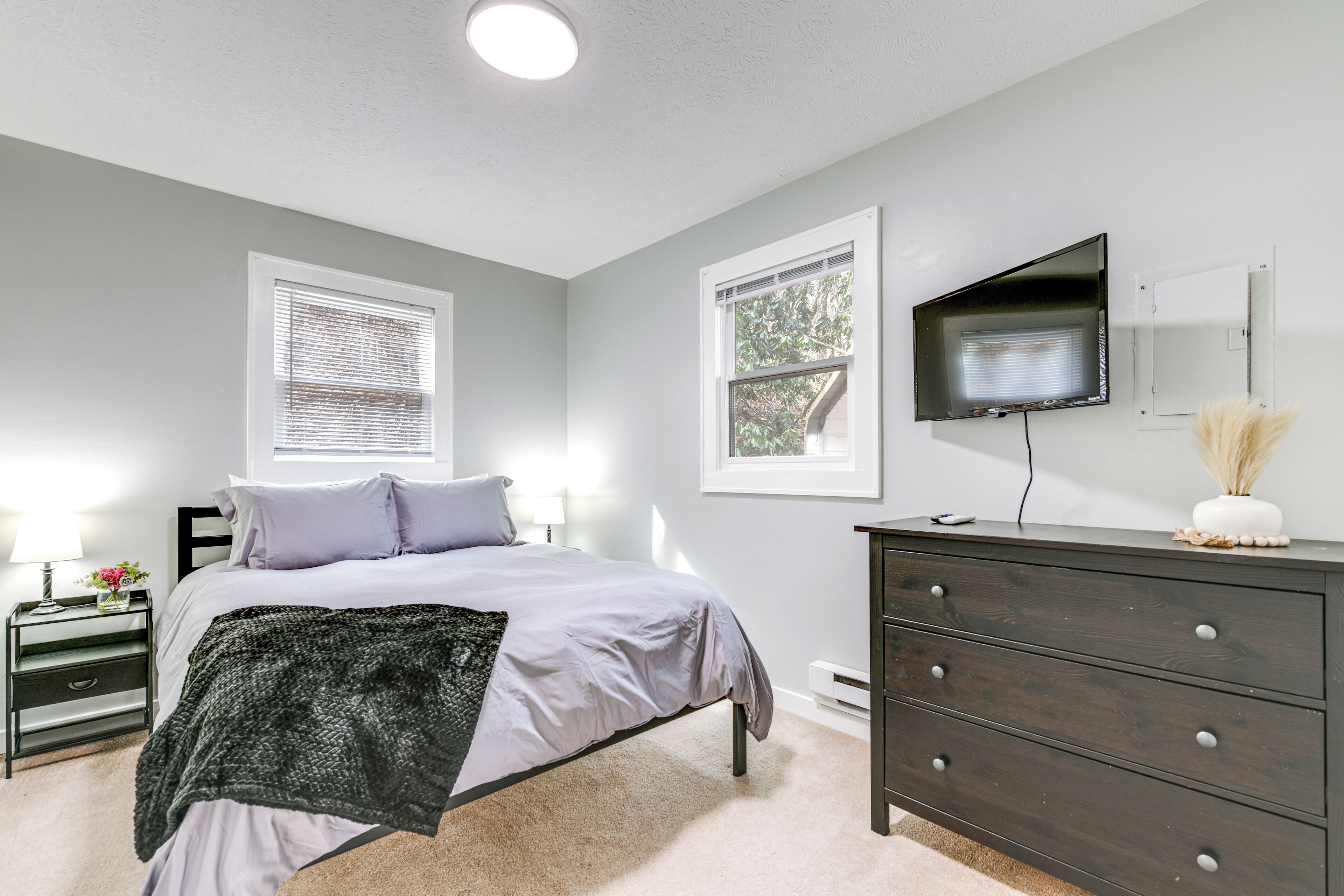 A bedroom with a bed, bedside tables, a dresser, a TV, and windows.