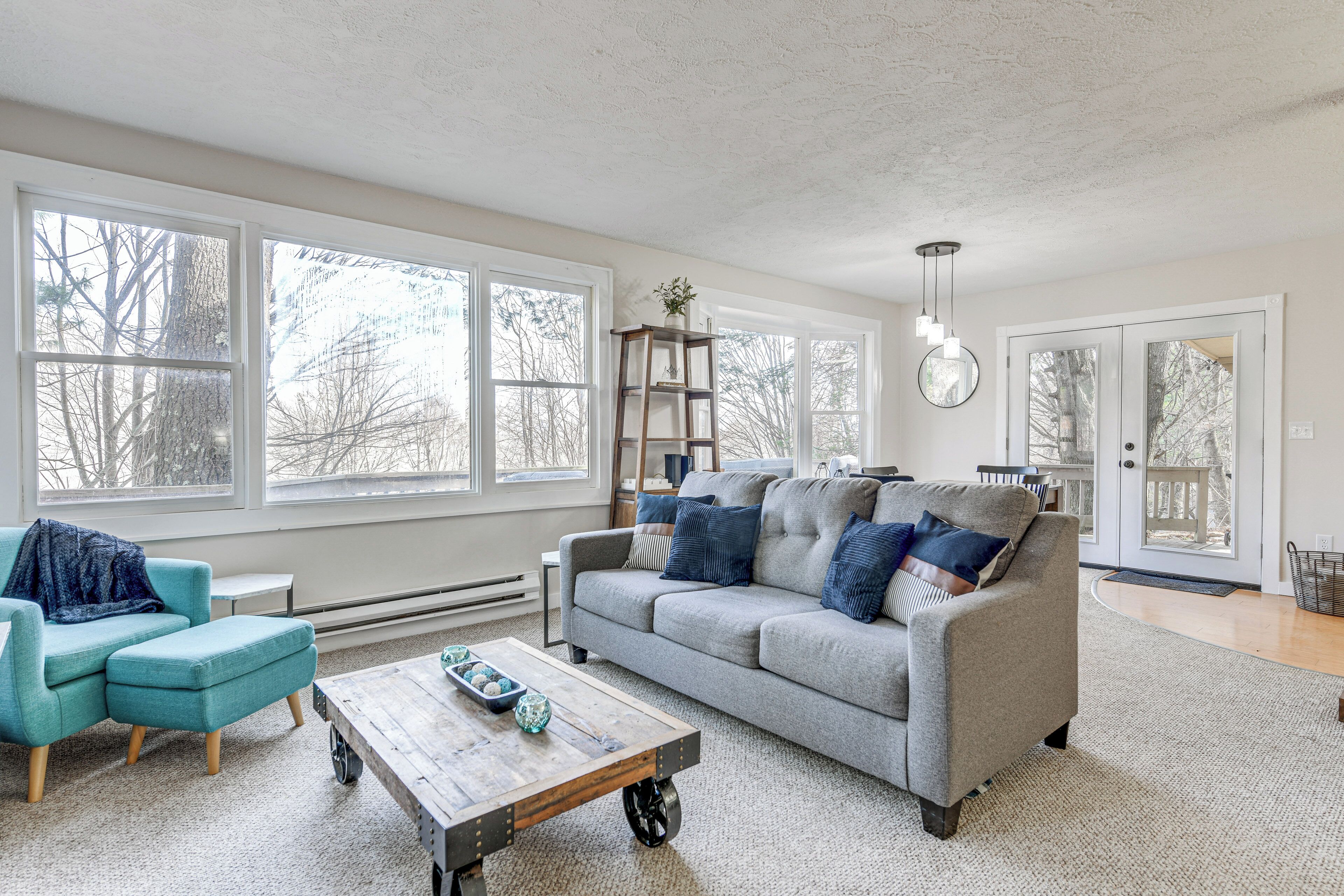 A spacious living room with large windows, a grey sofa with blue cushions, a wooden coffee table, and a small bookshelf.