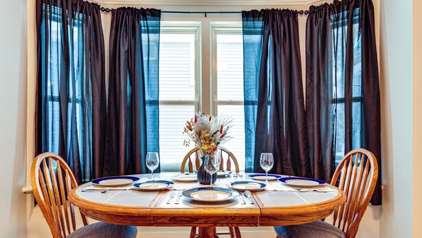 Una mesa de comedor dispuesta para cuatro personas, con sillas, un jarrón con flores y una ventana con cortinas.