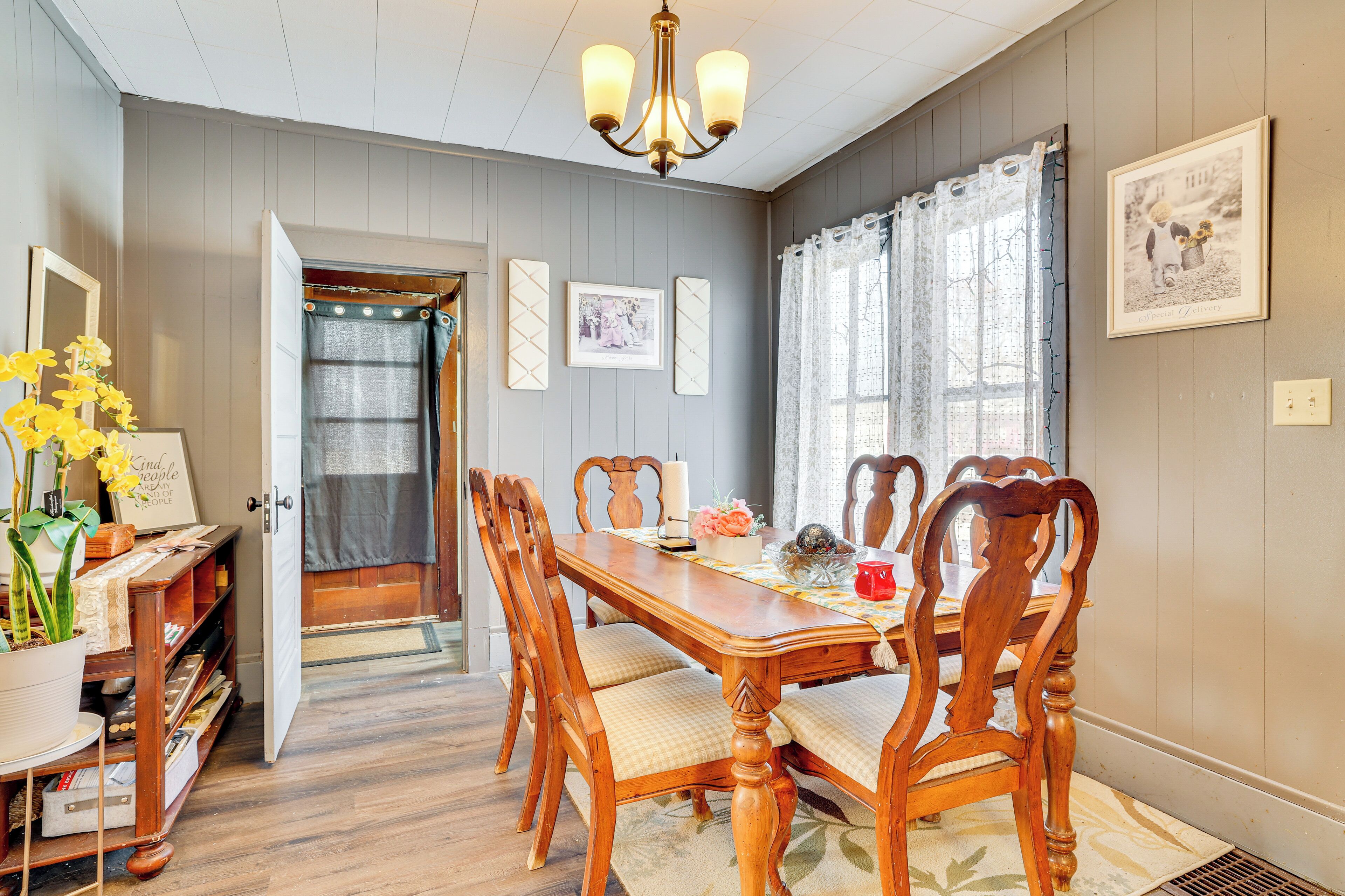 A dining room with a wooden table, chairs, and a chandelier. There is a door leading to another room, and a window with curtains.