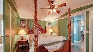 A four-poster bed with a wooden headboard, two bedside tables with lamps, a ceiling fan, and a view into an adjacent room.