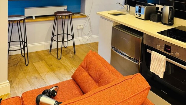 A compact kitchen with a built-in oven, a microwave, and a toaster. There is a window with blinds, a small bar stool, and a sofa with a hairdryer on it. - Leicester Square 2nd Floor Studio (London)
