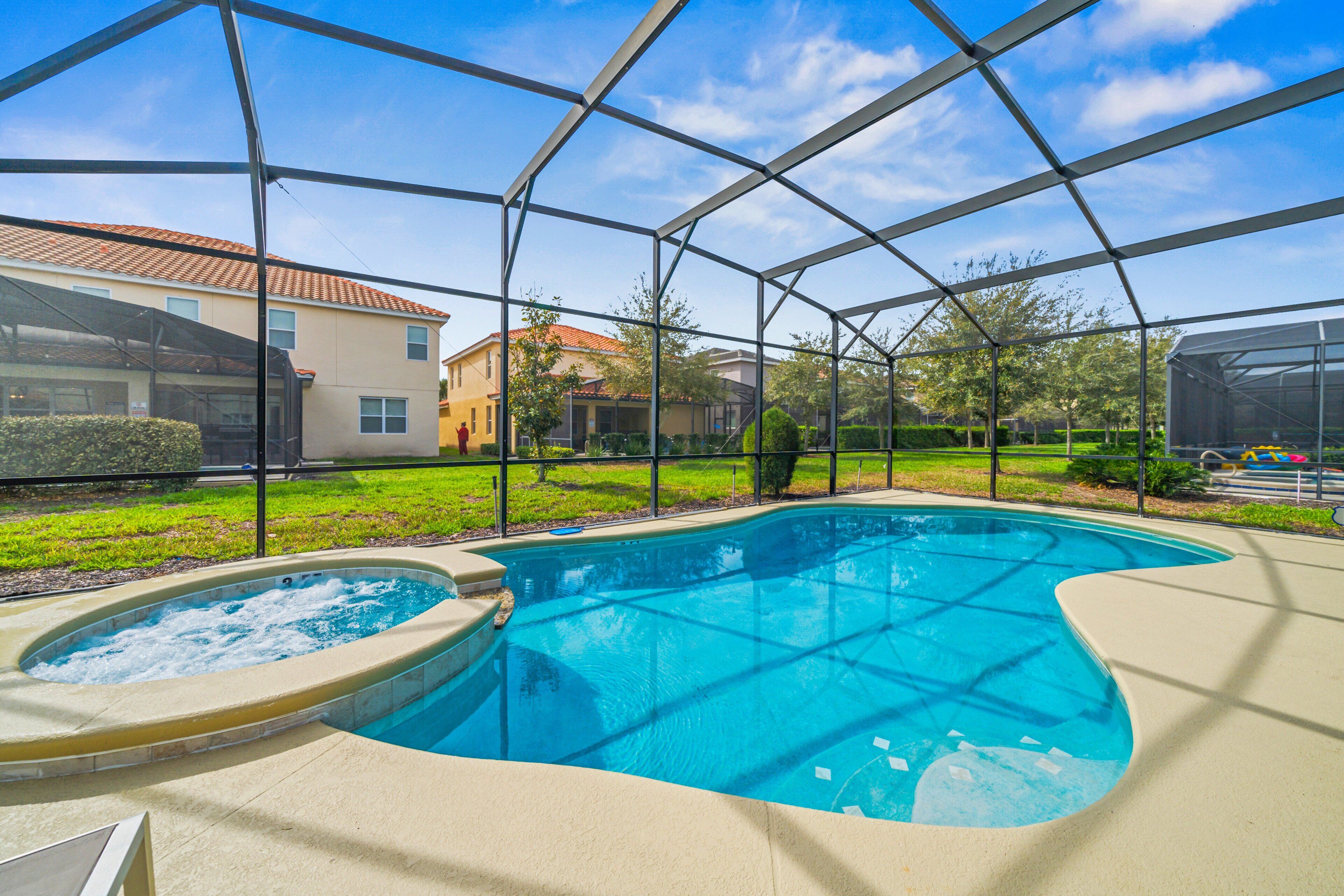 A swimming pool with a hot tub, surrounded by a covered area with a metal frame, and houses in the background.