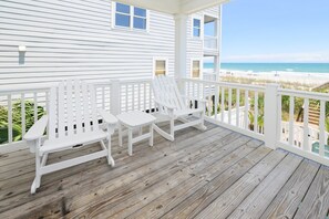 Una terraza de madera con sillas Adirondack blancas, barandilla y vista a la playa.