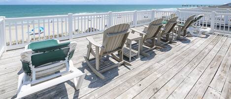 Une terrasse en bois avec des fauteuils Adirondack et un banc, donnant sur une plage avec une jetée.