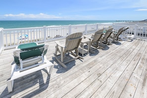 Una terraza de madera con sillas Adirondack y un banco, con vista a una playa y un muelle.