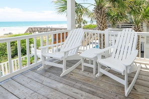 Una terraza de madera con sillas Adirondack blancas, una mesita y vista a la playa.