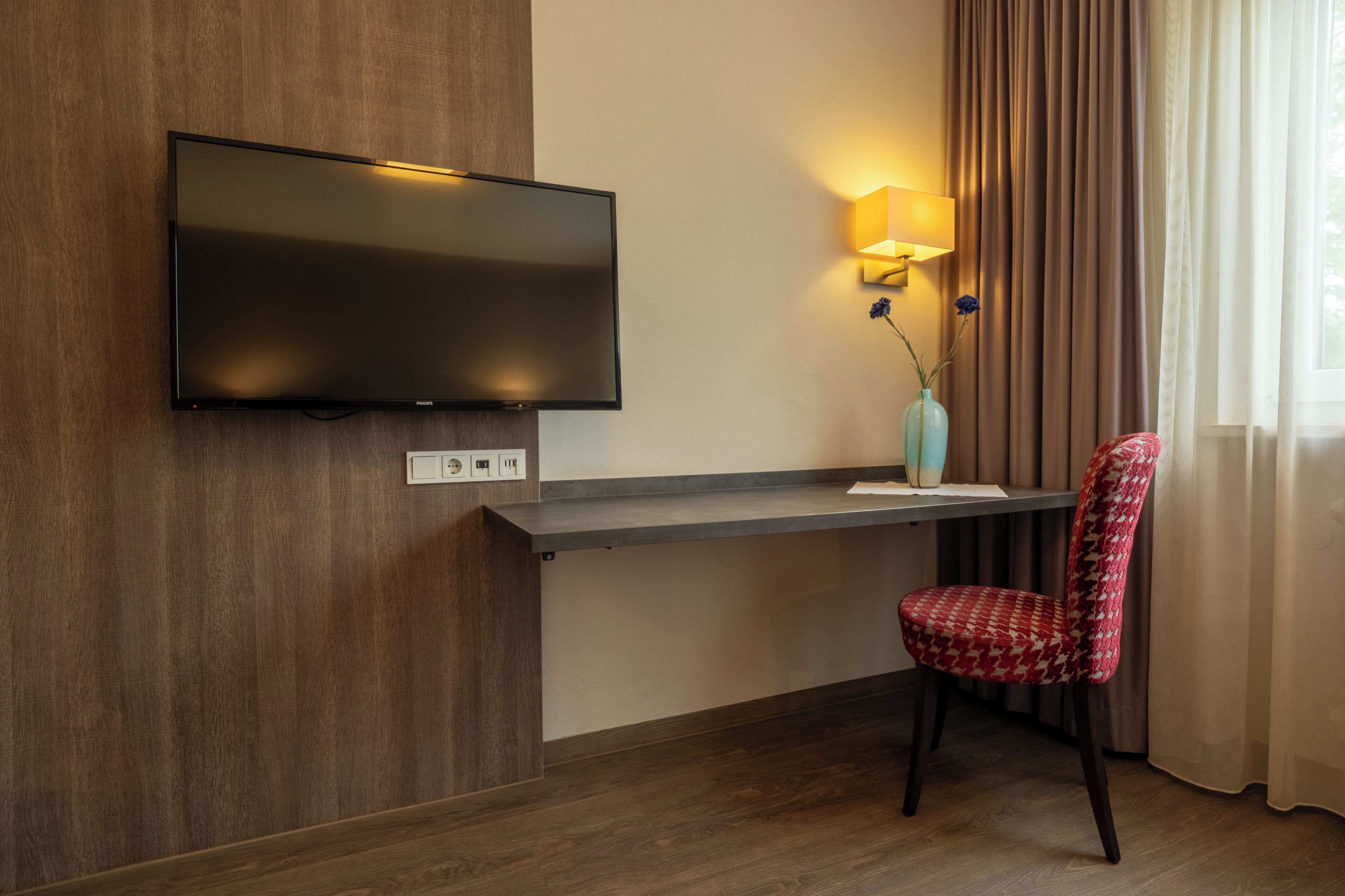 Une chambre d’hôtel moderne avec un téléviseur à écran plat fixé à un mur en bois, un bureau avec une lampe et une chaise à motifs.
