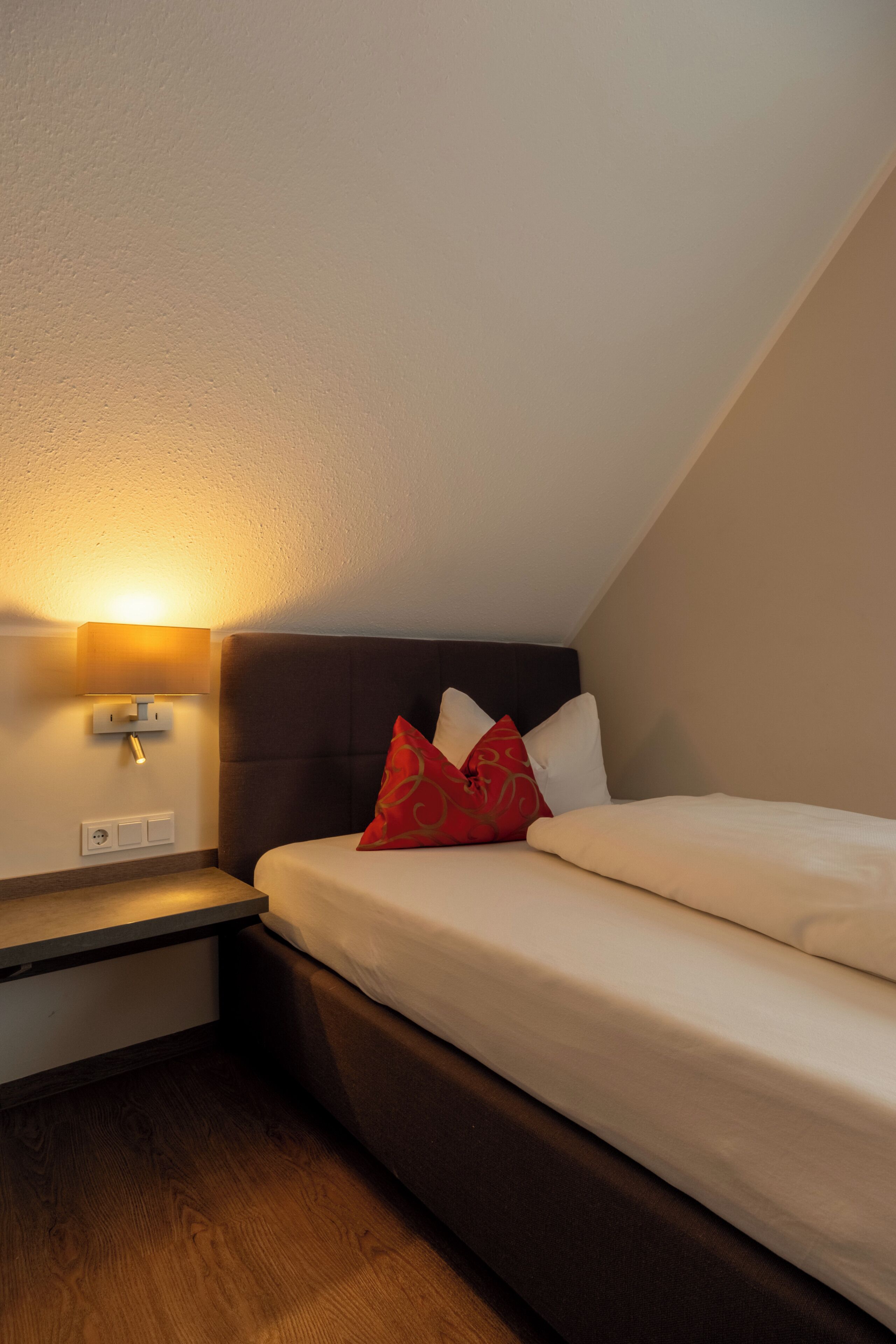 Een hotelkamer met een bed, een nachtkastje, een wandlamp en een houten vloer.