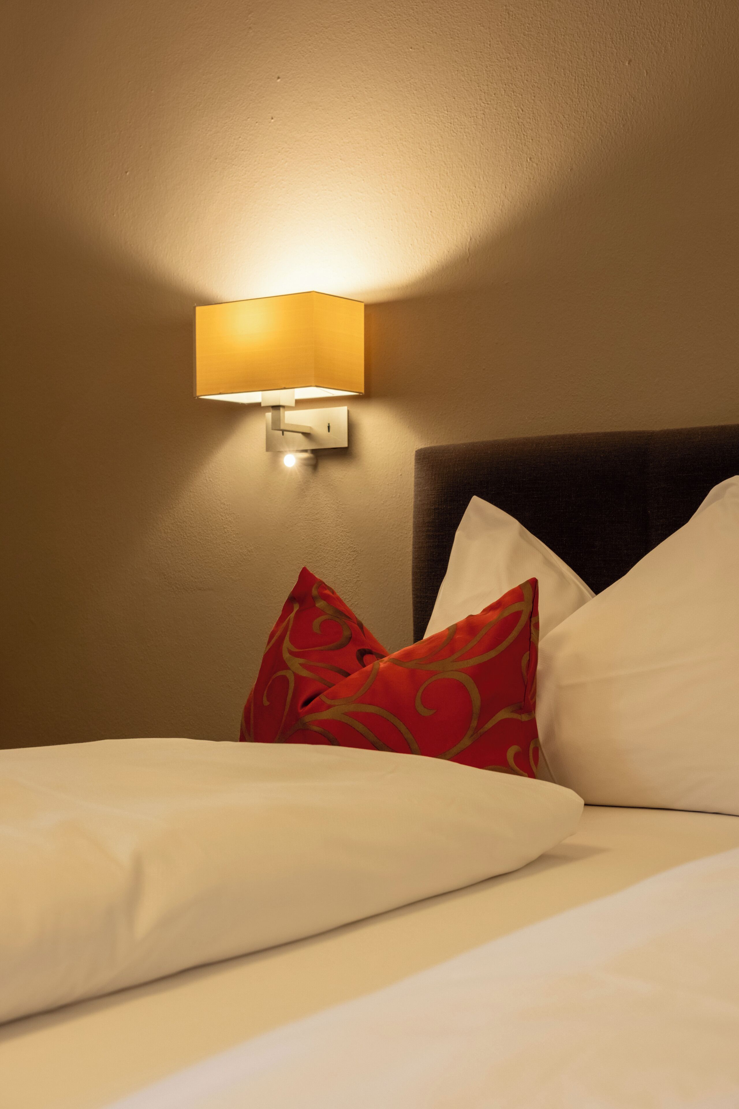 A hotel room with a bed, pillows, and a wall-mounted light fixture.