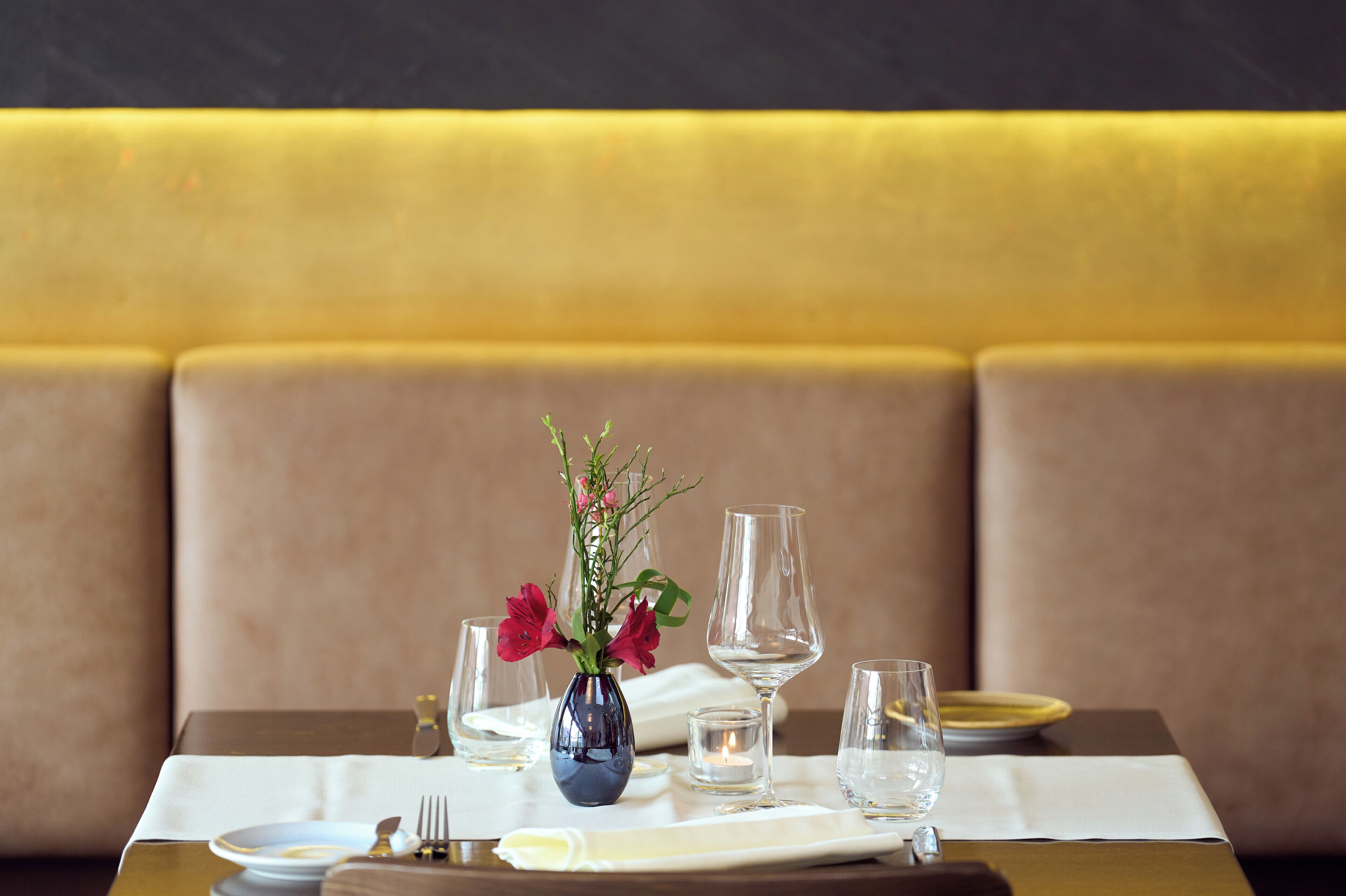 Una mesa de comedor dispuesta con un florero, copas de vino y platos, en una habitación con un banco acolchado.
