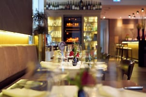 Un restaurante con zona de bar, mesas dispuestas con vasos y cubiertos, y un jarrón con flores sobre la mesa.