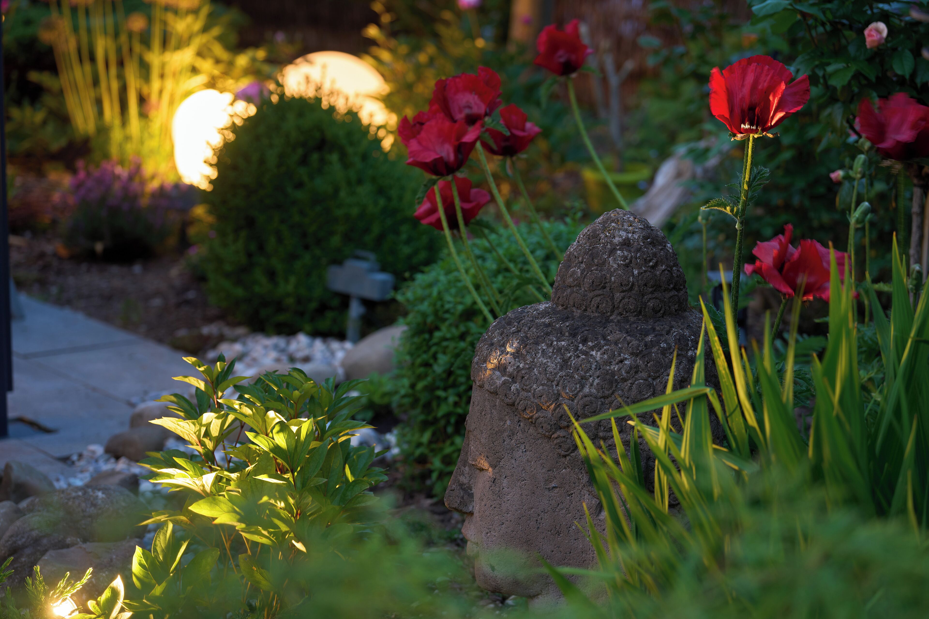Un jardín con una estatua de piedra, flores rojas y follaje verde.