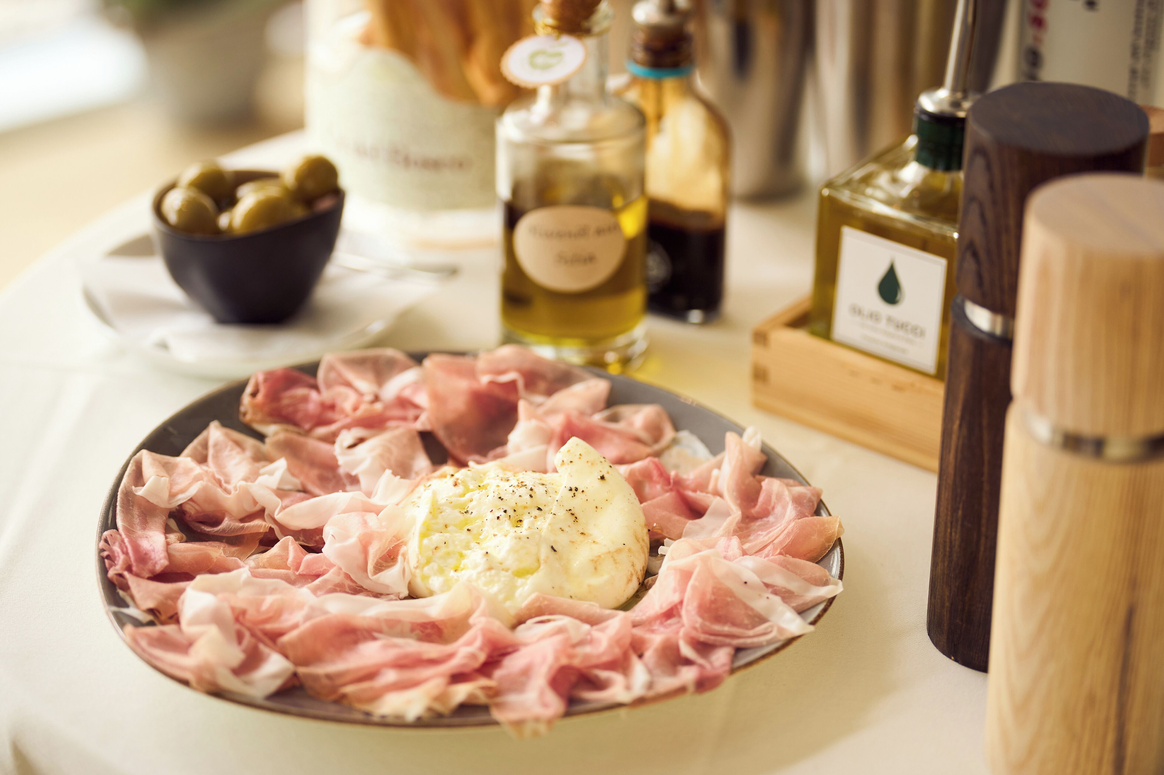 A plate of cured meats and cheese with a bottle of olive oil and a salt shaker on the table.