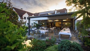 An outdoor dining area with tables and chairs, surrounded by greenery and a building with a tiled roof. - Hotel Staffler & Schreiners Restaurant (Odelzhausen)
