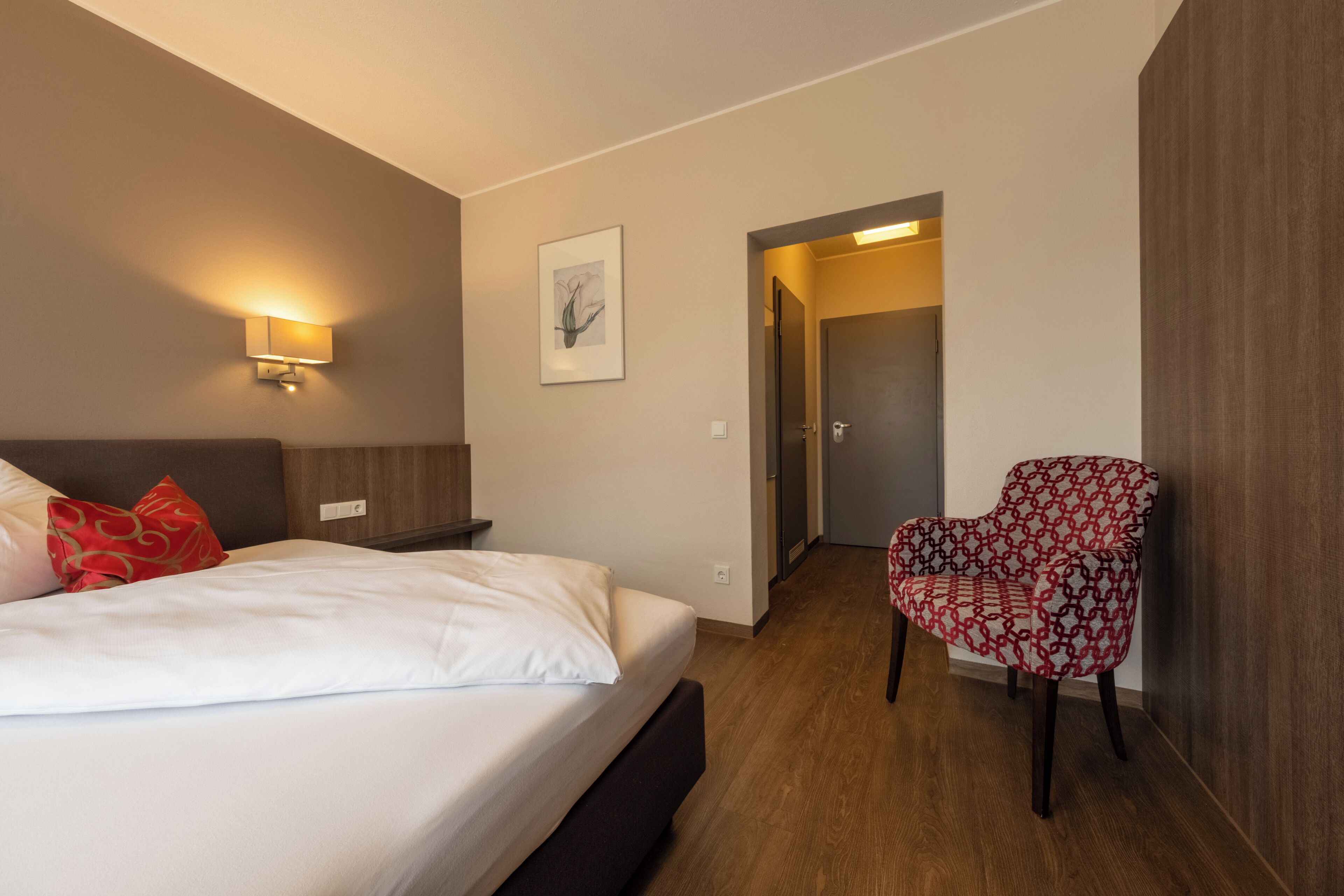 Habitación de hotel con cama, mesita de noche, lámpara, sillón y puerta que da a otra habitación.